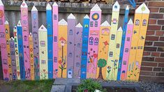 a wooden fence painted with colorful houses and trees in front of a brick wall next to a flower bed