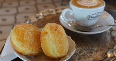 two sugary donuts on a plate next to a cup of cappuccino