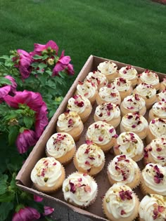 a box full of cupcakes with white frosting and pink flowers in the background