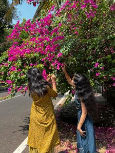 two women standing next to each other in front of purple flowers and trees on the street