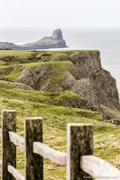 a wooden fence on top of a grass covered hillside next to the ocean with an island in the distance