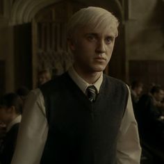 a man with white hair wearing a vest and tie in a room full of people