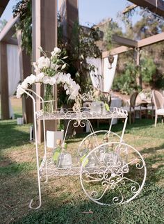 a white metal cart with flowers and glasses on it sitting in the grass near some chairs