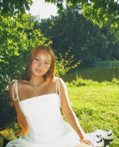 a woman in a white dress is sitting on the grass near some trees and water