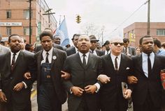 a group of men in suits and ties are walking down the street with people behind them