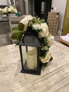 a black lantern with white flowers and greenery on the top sits on a wooden table