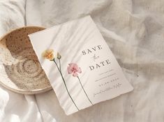 a wedding save the date card next to a bowl on a white cloth covered bed