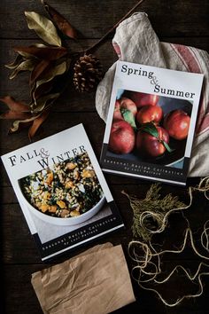 two cookbooks sitting on top of a wooden table next to some leaves and apples