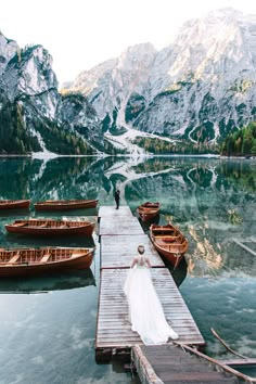 a bride and groom standing on a dock next to boats in the water near mountains