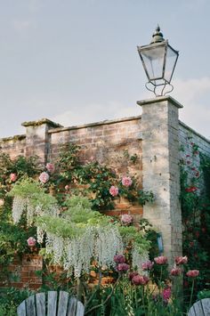 an old brick building with lots of flowers growing on it and two wooden chairs in the foreground