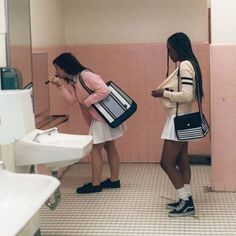 two women standing in a public restroom with their cell phones up to their ear and one holding a purse