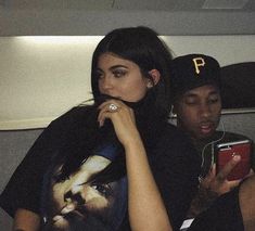 two people sitting next to each other while looking at their cell phones and one person wearing a baseball cap