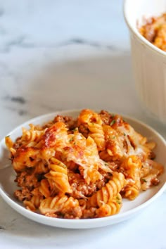 a white bowl filled with pasta and meat on top of a marble table next to a cup