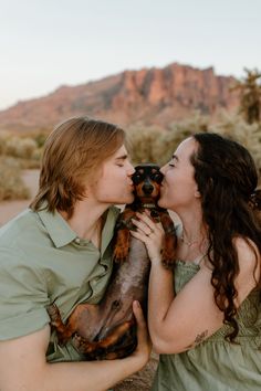 a man and woman kissing while holding a dog in the desert with mountains in the background