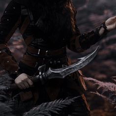 a woman dressed in black holding a large knife and looking at the camera while standing on rocks