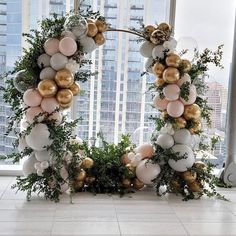 an arch made out of balloons and greenery in front of a window with skyscrapers