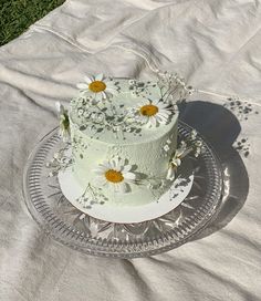 a white cake with daisies on it sitting on a glass plate in the grass