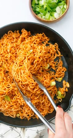 someone is holding tongs over some noodles in a skillet on the table next to a bowl of vegetables