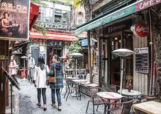 two women walking down an alley way in front of cafes and tables with umbrellas