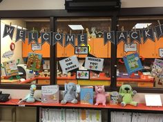 some books and stuffed animals are on a table in front of a window with welcome back signs