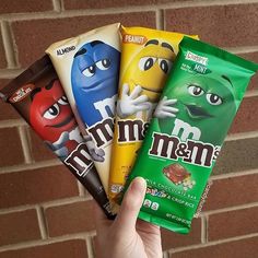 someone is holding four different flavors of m & m's candy bars in front of a brick wall