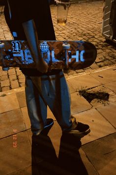 a man holding a skateboard on the sidewalk at night time with his feet in the air