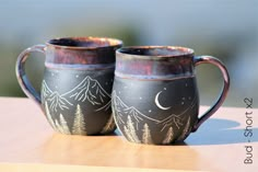 two mugs sitting on top of a wooden table with mountains and trees painted on them