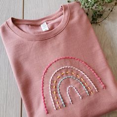 a pink t - shirt with an embroidered rainbow on the front, and a plant behind it