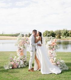 a bride and groom kissing in front of a floral arch on the grass by a lake