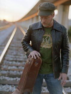 a man standing on train tracks holding a brown suitcase and looking at the ground with his hands in his pockets