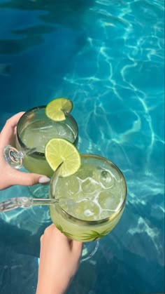 two people holding glasses with drinks in them near the water's edge and swimming pool