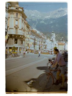 a man riding a bike down a street next to tall buildings with mountains in the background