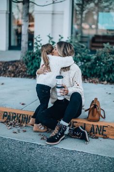 two women sitting on the curb kissing and hugging each other while holding a coffee cup