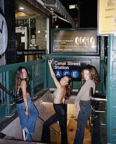 three girls are standing on the stairs in front of a subway station sign at night