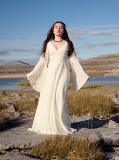 a woman in a long white dress standing on rocks near water and grass with her arms outstretched