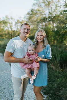 a man and woman holding a baby on a gravel road with trees in the background