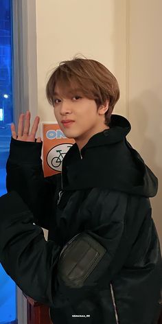 a young man standing in front of a window wearing a black jacket and holding his hand up