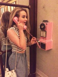 a woman talking on a pink phone while holding a purse and standing next to an old fashioned telephone