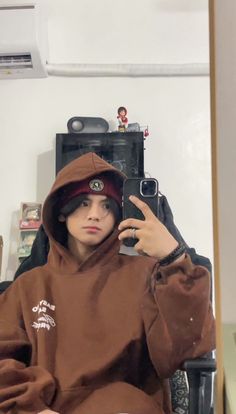 a person sitting in a chair taking a selfie with a cell phone and wearing a brown hoodie