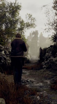 a man walking down a dirt road next to a stone wall covered in fog and trees