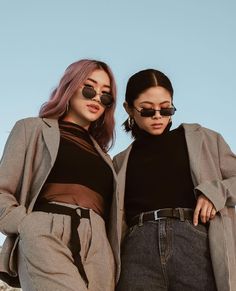 two women standing next to each other in front of a blue sky wearing black sunglasses