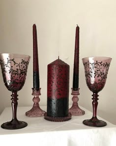 three candles and two wine goblets are sitting on a white tablecloth with a red candle in the middle