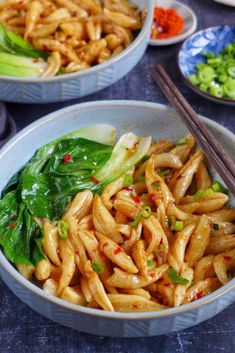 two bowls filled with noodles and vegetables next to chopsticks on a blue table cloth