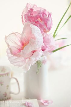 two pink flowers in a white vase on a table next to a cup and saucer