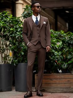 a man in a brown suit and sunglasses standing on the sidewalk with his hands in his pockets