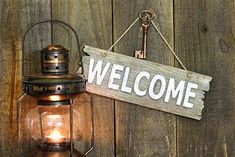 a sign that says welcome hanging on a wooden wall next to a lantern with a light