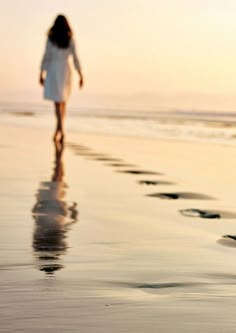 a woman walking on the beach with her reflection in the wet sand as the sun sets