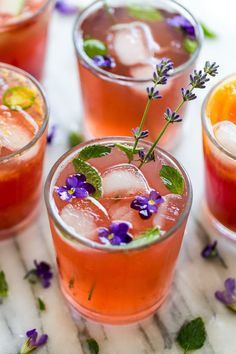 four glasses filled with different types of drinks and garnished with purple flowers on a marble surface