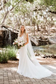 a woman in a wedding dress holding a bouquet and standing next to a river with trees