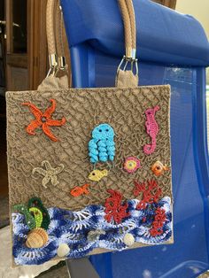 a handbag with crocheted sea creatures on the front and sides, hanging from a blue chair